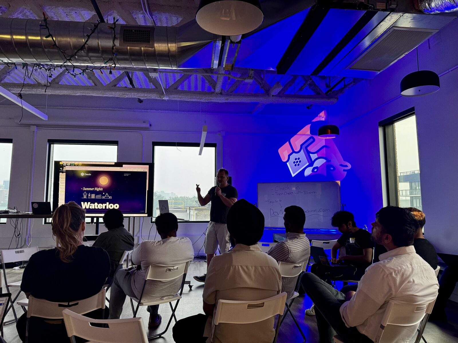 A speaker presenting to the seated room under blue light, a Summer Nights Waterloo slide on screen